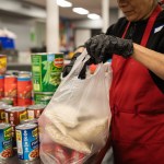 A person wearing a red apron and black gloves packs rice and canned goods into a plastic bag at a food distribution center. Rows of canned beans and vegetables are visible on the counter.