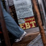 A person walks into a corner store displaying a large sign in the window that reads “We Accept EBT.”
