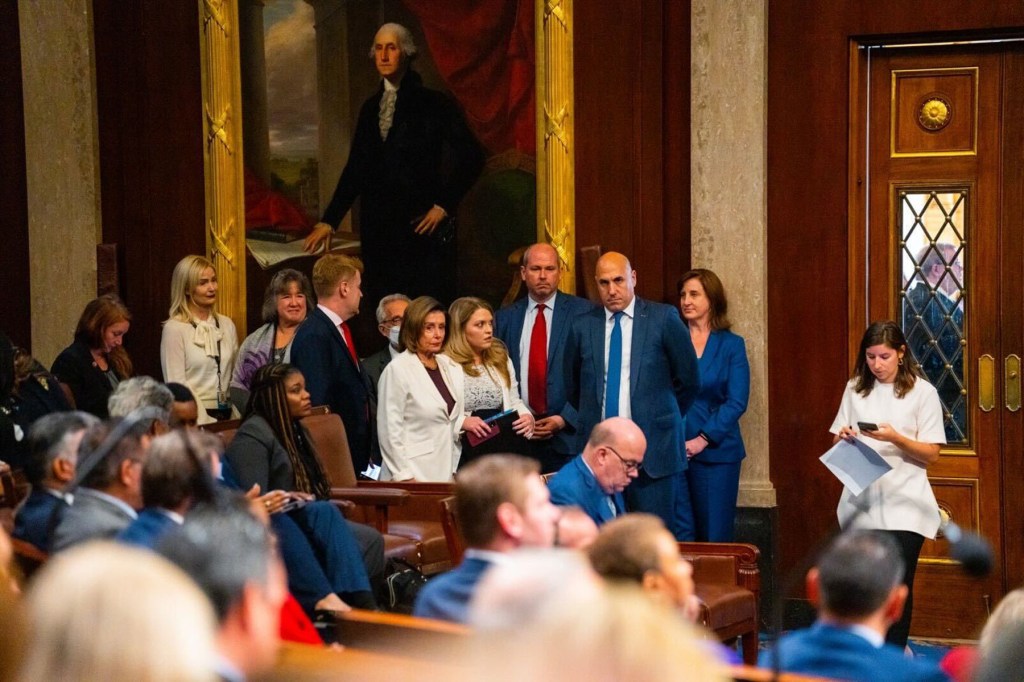 Members of Congress and staff stand in the U.S. House chamber near a large portrait of George Washington. Nancy Pelosi stands near the center of the group wearing a white suit, surrounded by aides and lawmakers. People are seated in the foreground as the chamber fills with members.