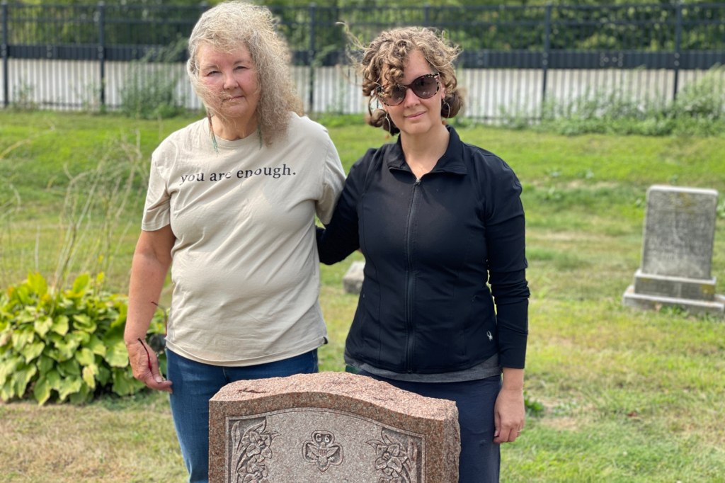 Two women stand at a gray headstone for Mary Dainty.