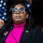 McIver stands at a mic in a pink shirt and black blazer flanked by two male politicians behind her.