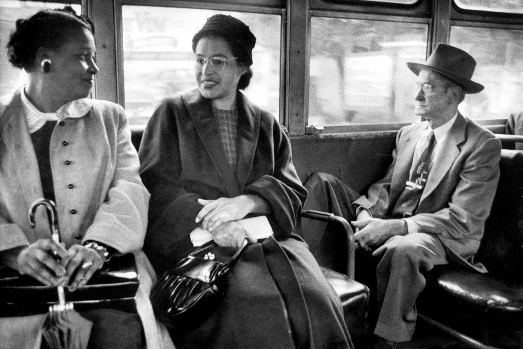 Two women smile and talk seated on a bus alongside a man looking out the window.