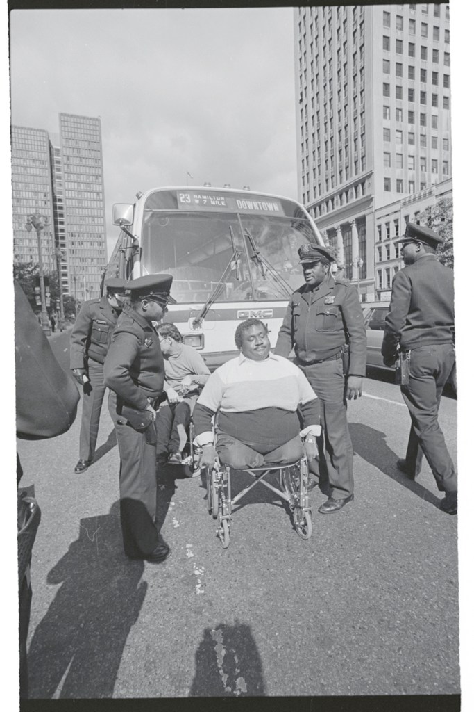 A man using a wheelchair is in front of a city bus surrounded by police officers.
