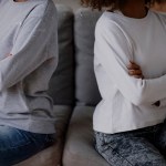 A mom and a teen sit side by side on a couch with their arms crossed, facing away from each other.