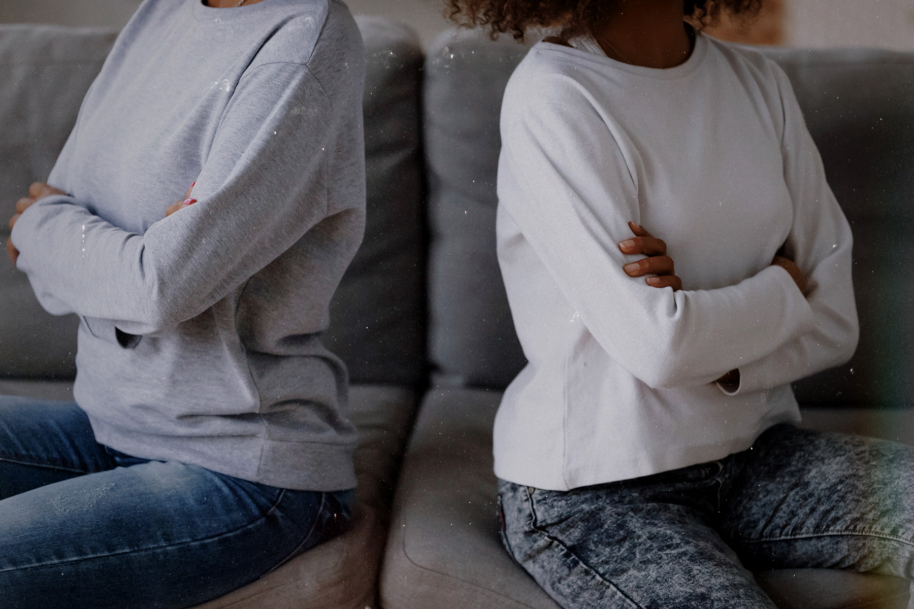 A mom and a teen sit side by side on a couch with their arms crossed, facing away from each other.