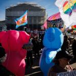 Protesters gather in front of the Supreme Court. Two people wearing inflatable costumes are in the foreground, and flags with marriage equality symbols are being waved.