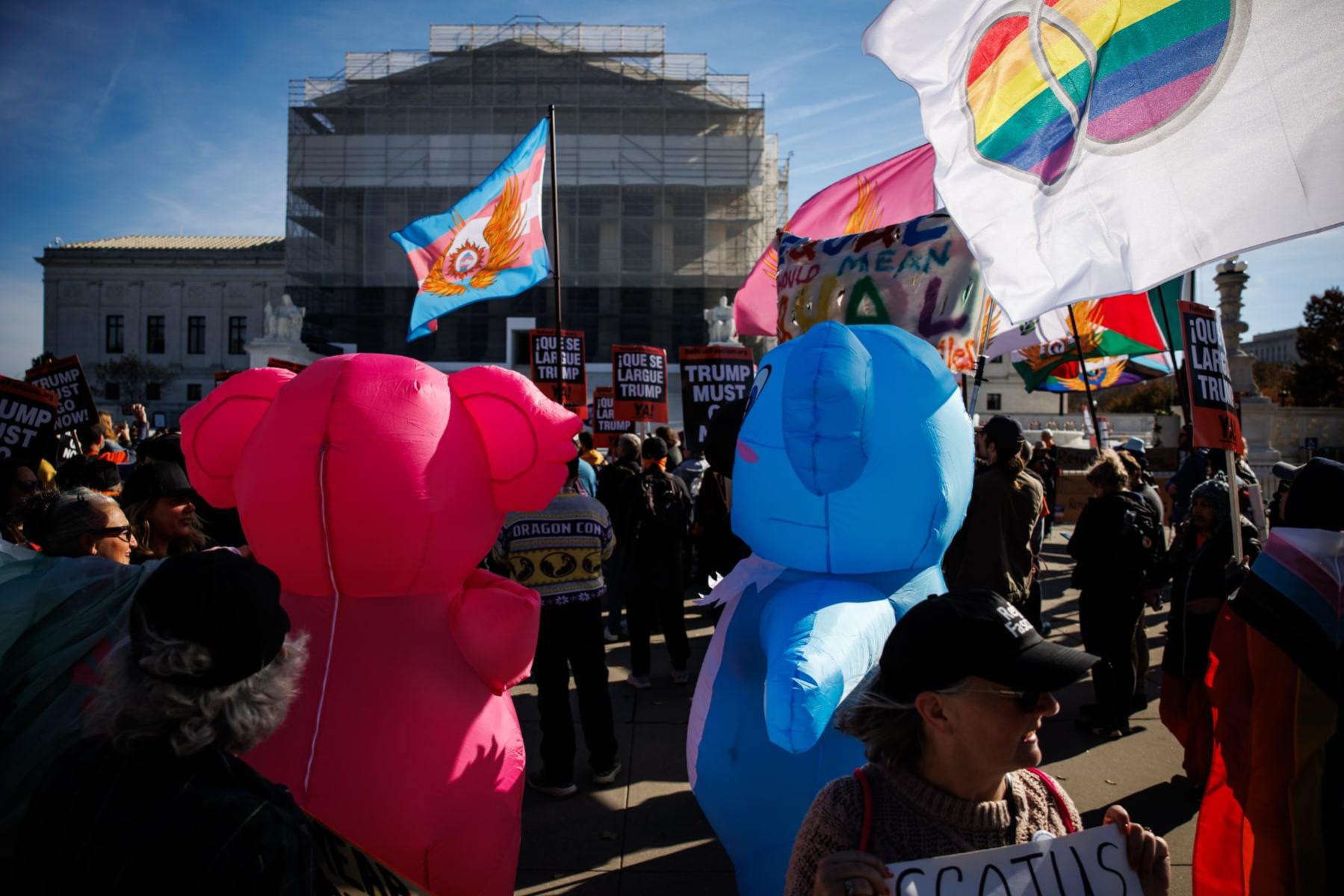 Protesters gather in front of the Supreme Court. Two people wearing inflatable costumes are in the foreground, and flags with marriage equality symbols are being waved.