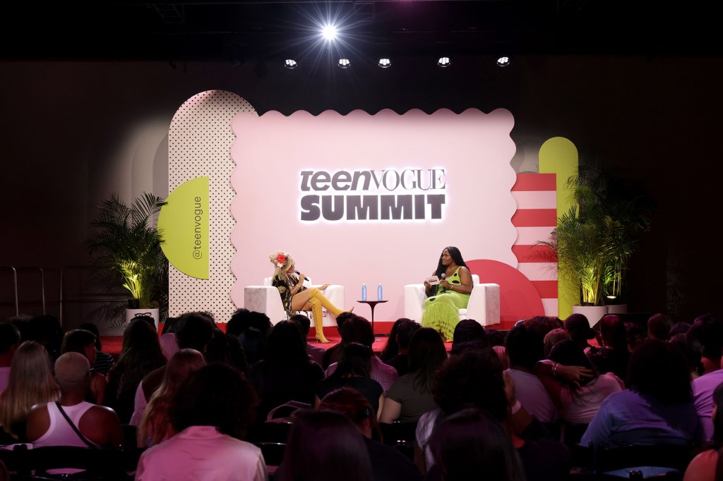 A speaker and a musician sit onstage in white armchairs at the Teen Vogue Summit, holding microphones as they talk. The stage backdrop reads “Teen Vogue Summit” and colorful graphic shapes and plants surround the set. An audience fills the foreground.