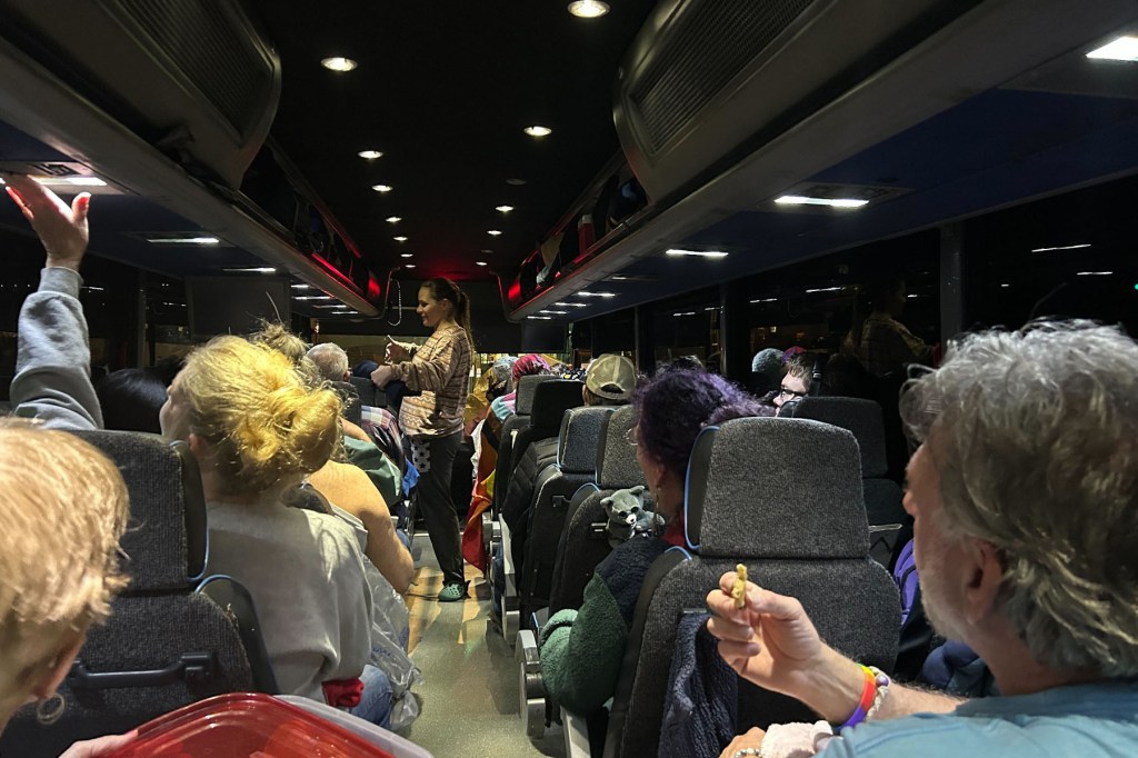 A crowded charter bus at night with passengers sitting closely together in gray seats. Some people hold snacks and containers of food, and others adjust overhead lights or settle in with blankets. A woman stands in the aisle.