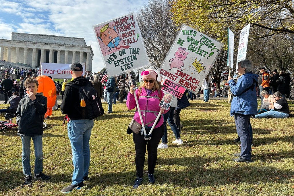 Judy Routhier holds two signs that read, “Hump-y Trump- Must Fall! He’s on the Epstein List” and “This ‘Piggy’ Won’t be Quiet! IMPEACH. CONVICT. REMOVE.”