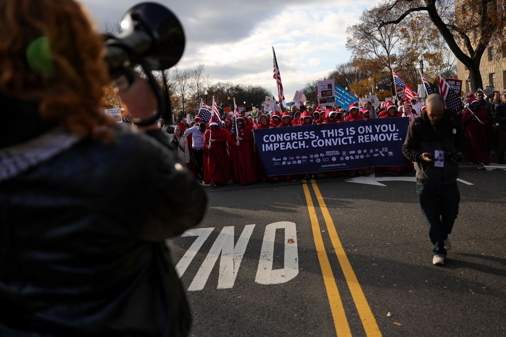 Protesters in red Handmaid’s Tale cloaks join the march in Washington, D.C., carrying a large banner calling on Congress to “Impeach. Convict. Remove.”