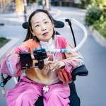Alice Wong, using a power wheelchair, sits outdoors on a paved path. She has a tracheostomy tube connected to a ventilator and holds her hands together near the controls mounted in front of her. She wears a multicolored long-sleeve top and bright pink pants.