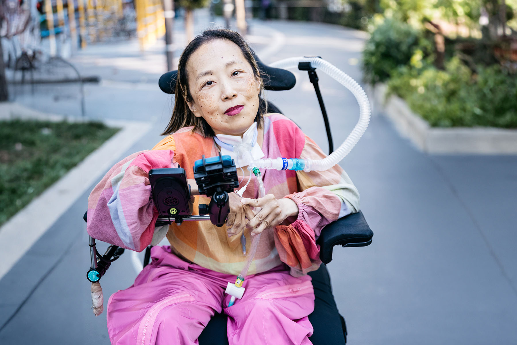 Alice Wong, using a power wheelchair, sits outdoors on a paved path. She has a tracheostomy tube connected to a ventilator and holds her hands together near the controls mounted in front of her. She wears a multicolored long-sleeve top and bright pink pants.