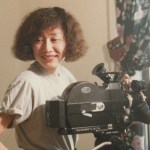 Filmmaker Christine Choy behind the scenes making a film, standing beside a film camera.