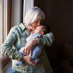 A grandmother wearing a plaid shirt holds a baby in a blue onesie by a window.