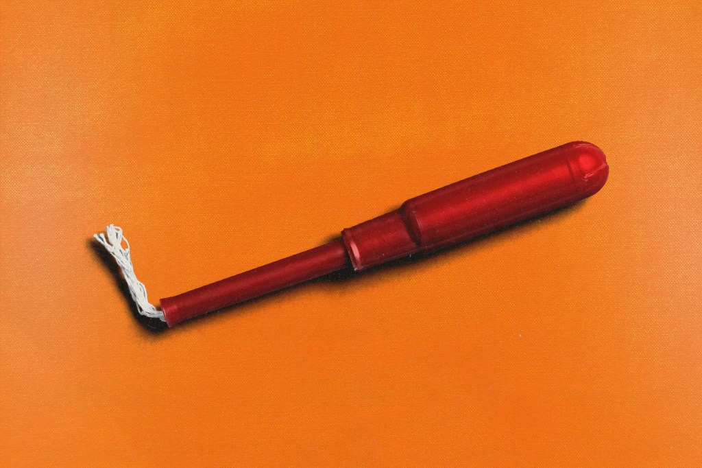 A red tampon with a frayed white string lying diagonally on a bright orange surface.