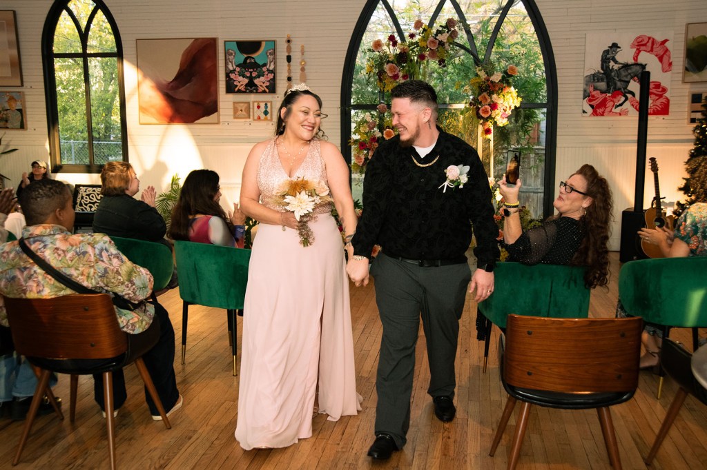 A newly married couple walks up the aisle holding hands and smiling at each other. Guests seated on green chairs clap as the pair passes the floral altar inside the bright, art-filled venue.