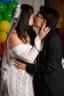 A close-up of a couple kissing, the bride in an off-shoulder lace gown and veil holding her partner’s face. They stand beside a backdrop of rainbow-colored balloons.
