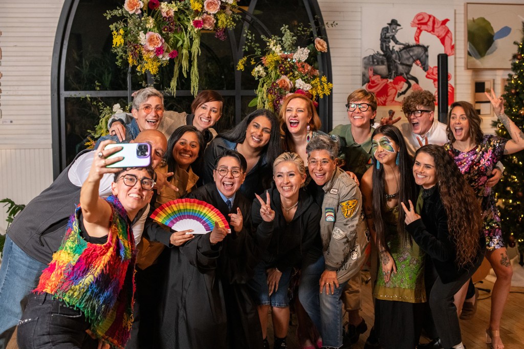 A large group of attendees gathers for a selfie in front of a floral arch inside the venue. People smile, pose, and gesture toward the camera.