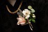 A close-up of a pale pink and white boutonnière pinned to a black patterned shirt, with a gold chain visible around the wearer’s neck.
