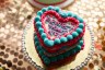 A heart-shaped cake decorated in pink and teal frosting sits on a gold cake board. The top reads “Protect Trans Kids.”
