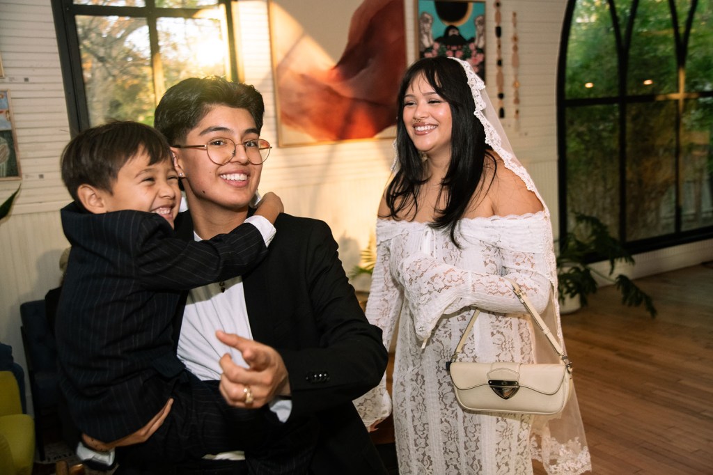 A person in a suit holds a laughing young child in their arms while the bride in a lace gown walks beside them, smiling inside the venue.