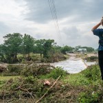A person stands on a grassy embankment looking out over flood damage after a severe storm, with downed trees, debris, and fast-moving water near a low bridge.