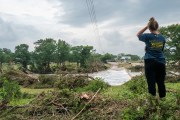 A person stands on a grassy embankment looking out over flood damage after a severe storm, with downed trees, debris, and fast-moving water near a low bridge.