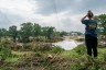 A person stands on a grassy embankment looking out over flood damage after a severe storm, with downed trees, debris, and fast-moving water near a low bridge.