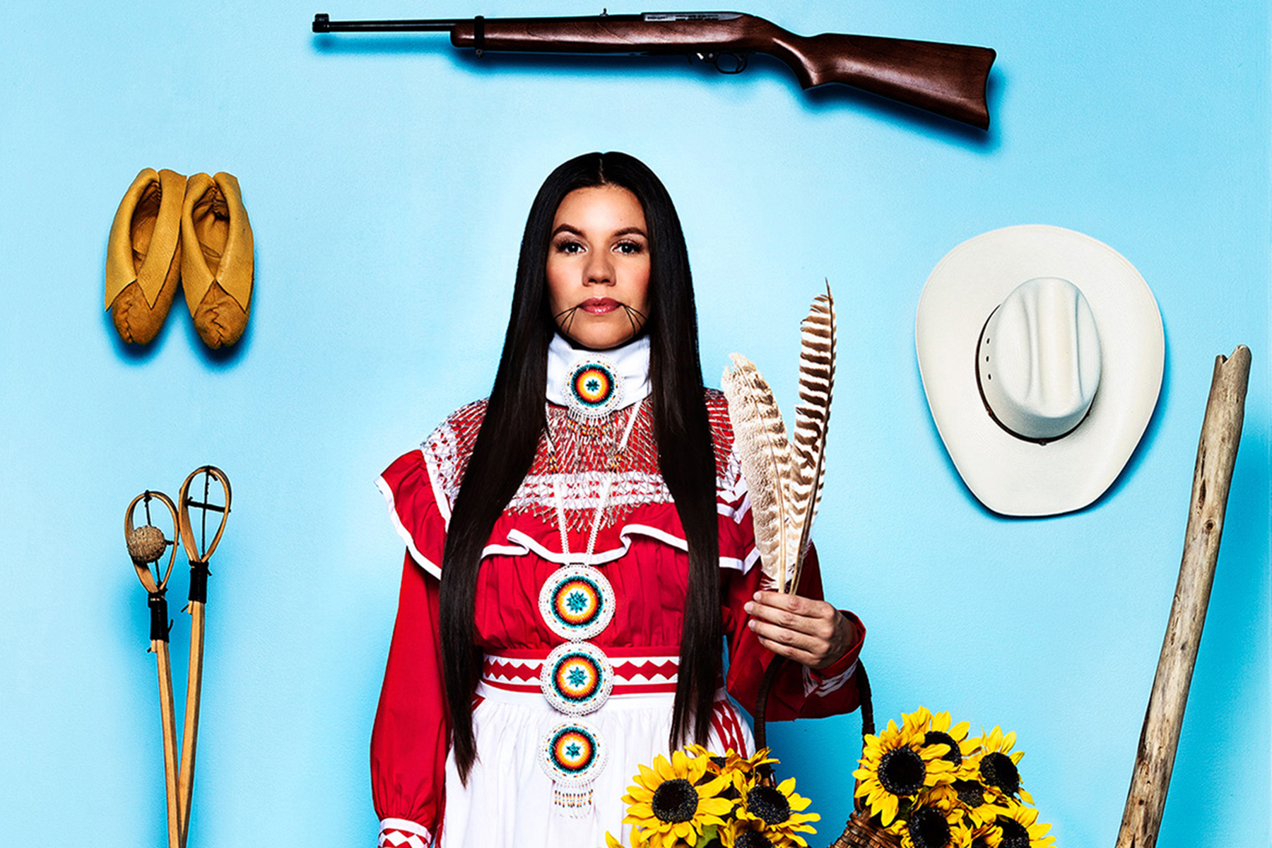 An image of an Indigenous woman wearing and surrounded by symbols of her tribal culture, photographed within a life-size box made to look like a doll’s box.