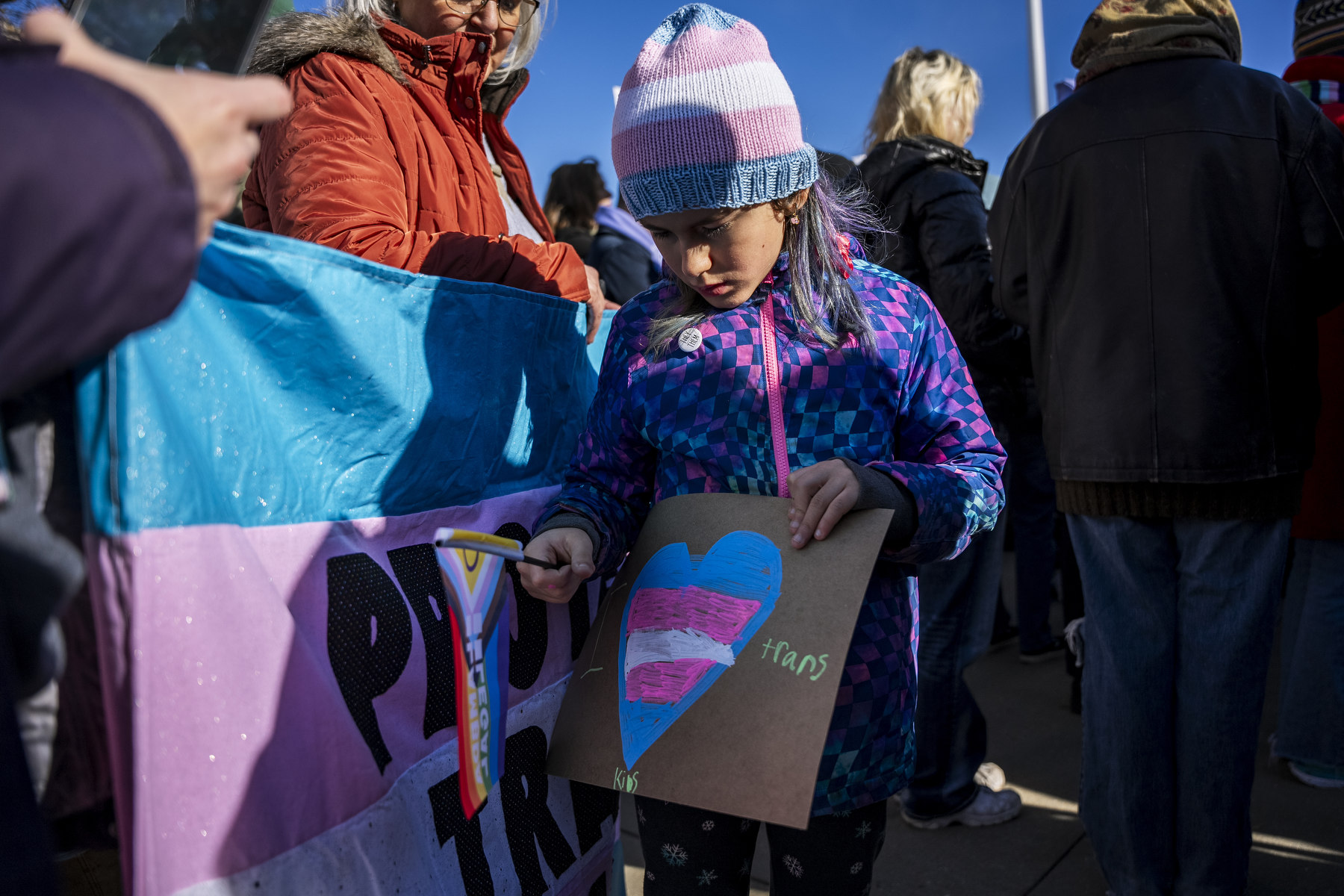 A child holds a sign with a heart in the colors of the trans pride flag.