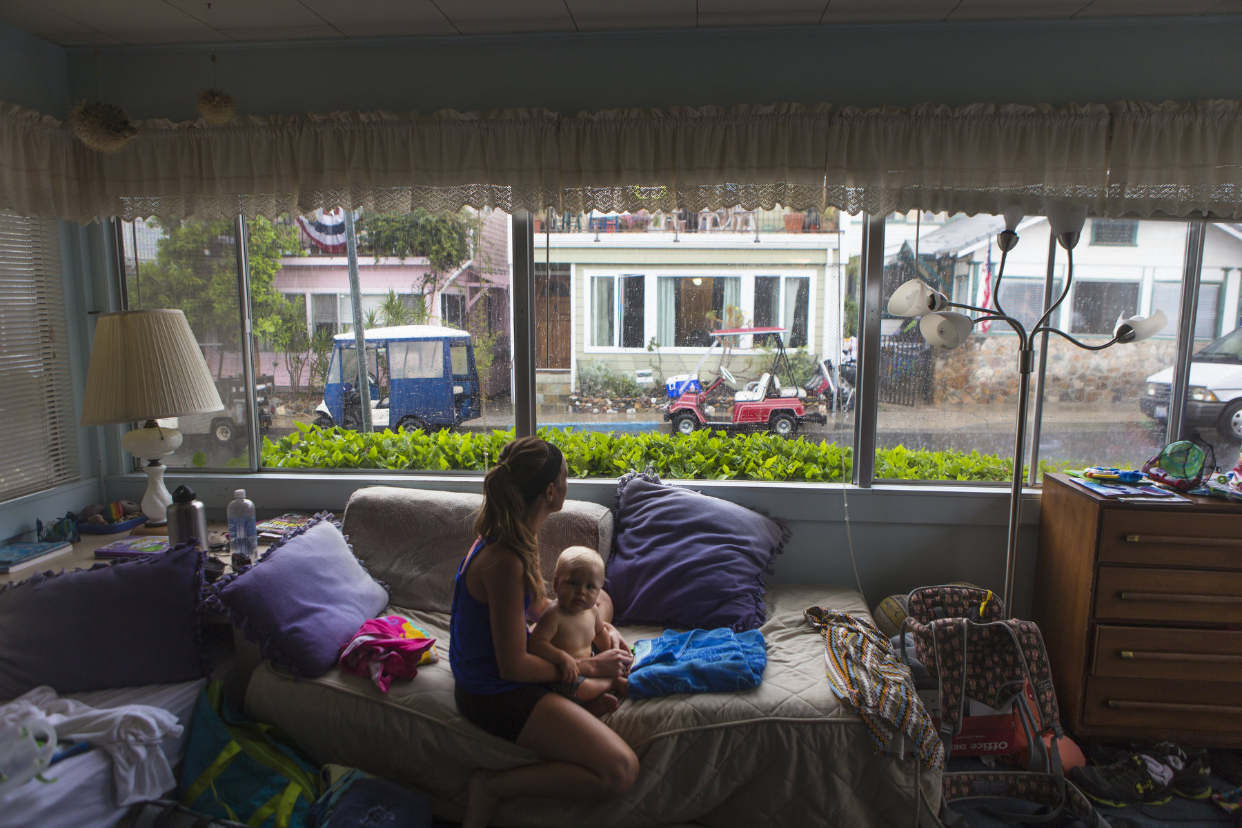 A woman holds a baby in a messy home as she looks out the window.