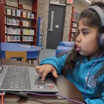 A young girl sits at a laptop in a library with headphones on.