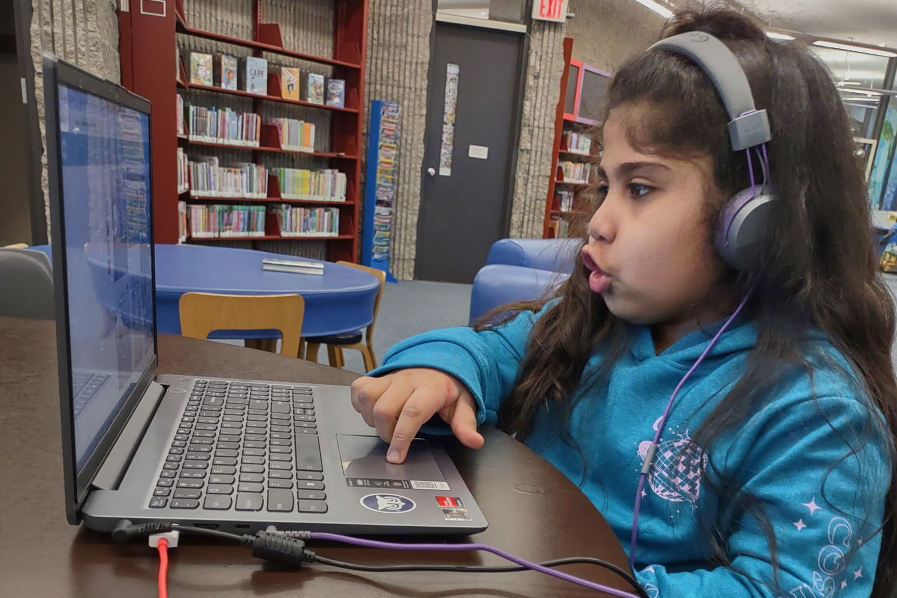 A young girl sits at a laptop in a library with headphones on.