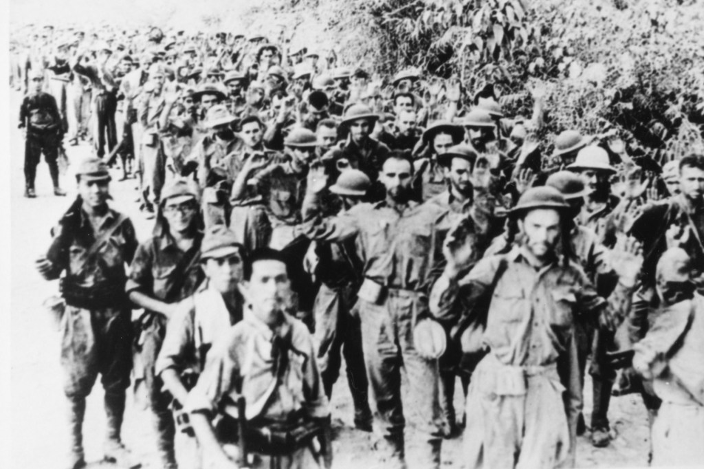 A long line of soldiers walk through the jungle in a black and white photo.