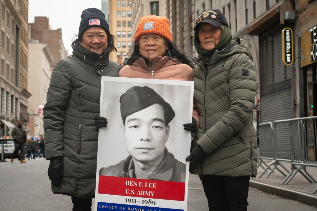 Three women hold a poster showing their grandfather and American flags on a street.