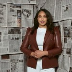 A woman stands facing the camera in front of a wall covered with newspaper pages.