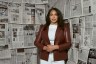 A woman stands facing the camera in front of a wall covered with newspaper pages.
