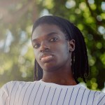 A young Black man stands outdoors beside a tree, looking directly at the camera. Sunlight filters through leaves in the background.