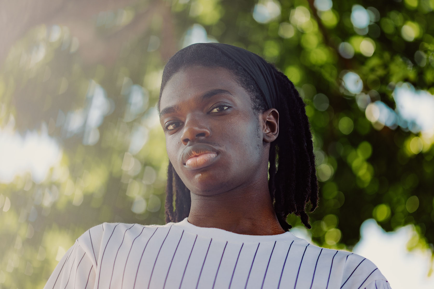 A young Black man stands outdoors beside a tree, looking directly at the camera. Sunlight filters through leaves in the background.