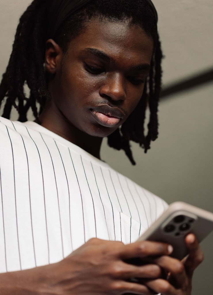 A young Black man looks down at a smartphone he holds in both hands. The image is tightly framed on his upper body.