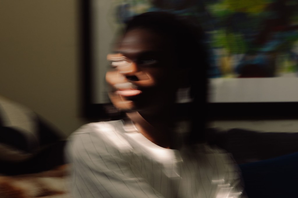 A blurred image of a young Black man seated indoors, his face partially obscured by motion. Light enters from the side of the frame.