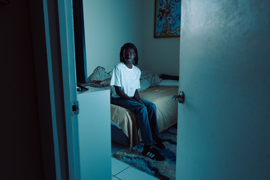A young Black man sits on the edge of a bed, seen through a partially open door. The room is dimly lit with blue-toned light.