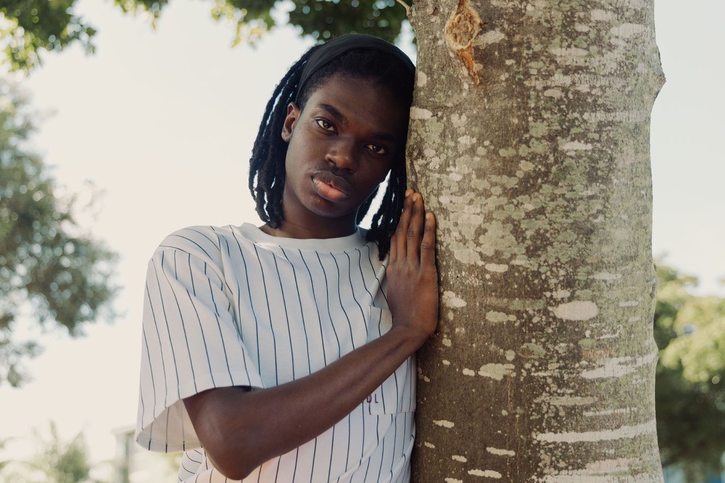 A young Black man stands outdoors beside a tree, resting one hand against the trunk. Greenery fills the background.