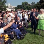President George Bush and First Lady Barbara Bush walk across a grassy lawn, waving to a seated crowd of people with disabilities, many using wheelchairs, during a public ceremony at the White House.