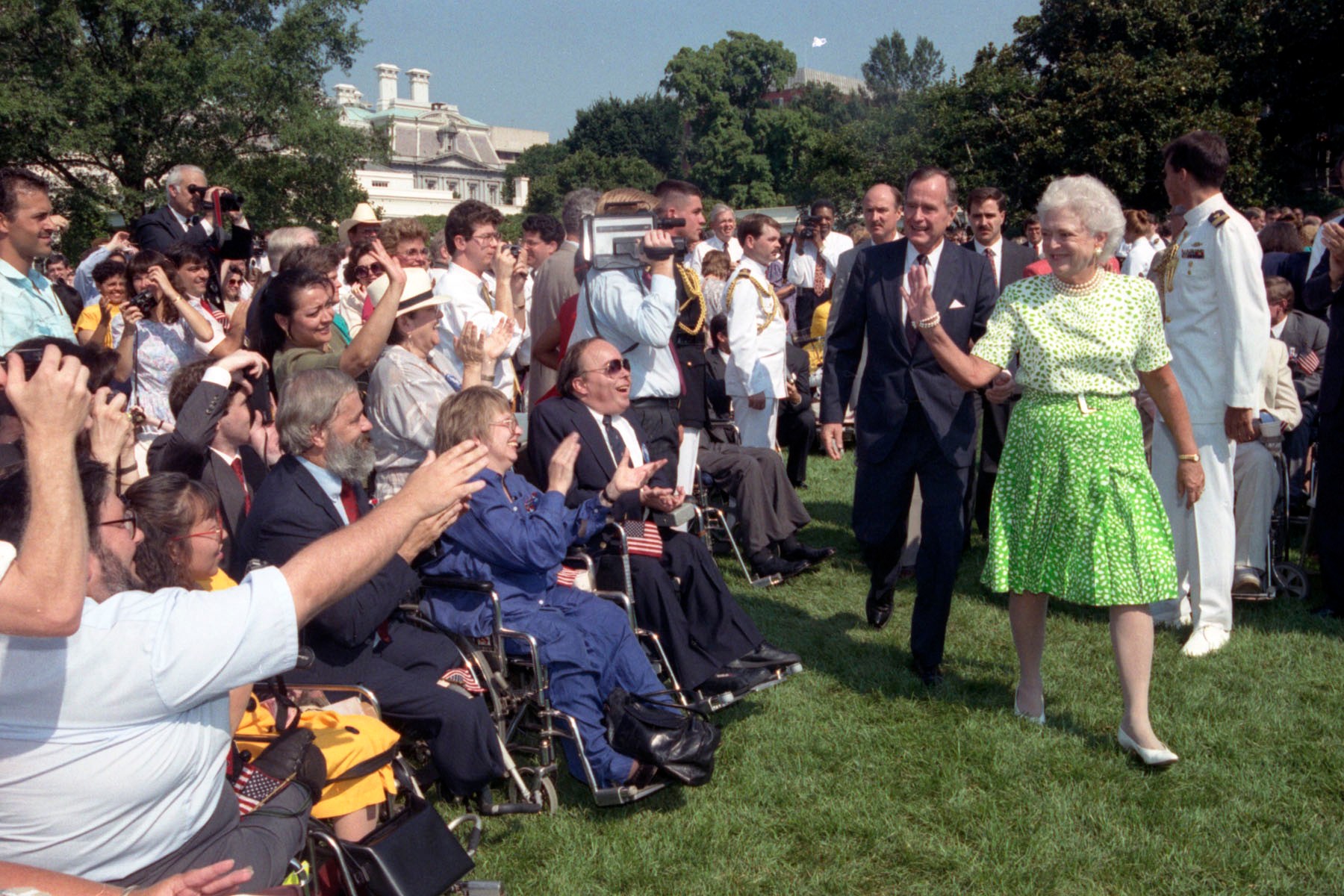 President George Bush and First Lady Barbara Bush walk across a grassy lawn, waving to a seated crowd of people with disabilities, many using wheelchairs, during a public ceremony at the White House.
