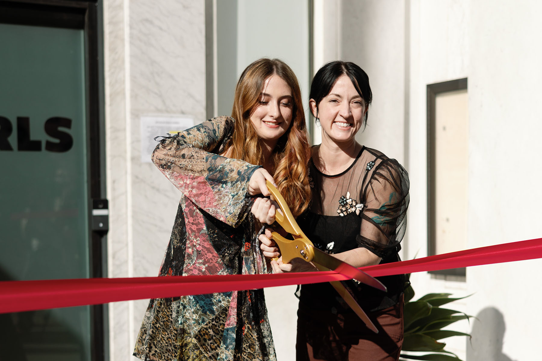 A teenage girl and an adult woman smile as they cut a red ribbon with oversized gold scissors outside a community space.