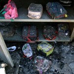 Children’s backpacks and lunch bags sit inside a metal storage cabinet, coated in ash and soot, with debris scattered across the floor after apparent fire damage.