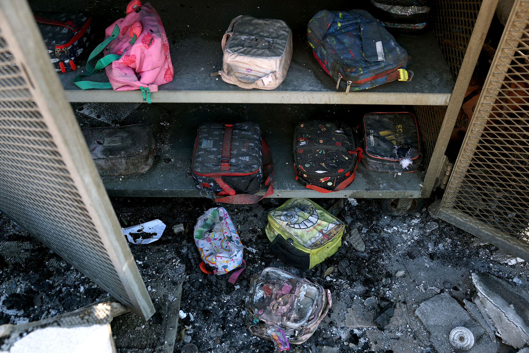 Children’s backpacks and lunch bags sit inside a metal storage cabinet, coated in ash and soot, with debris scattered across the floor after apparent fire damage.