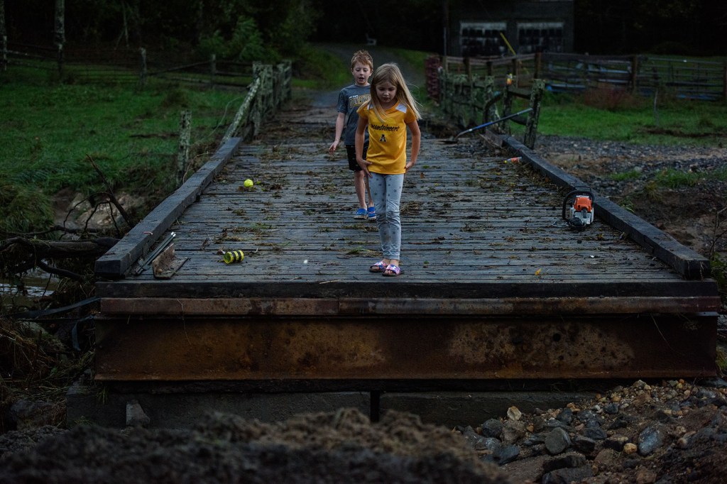 Two young children walk across a damaged wooden bridge littered with debris, including tools and broken boards.